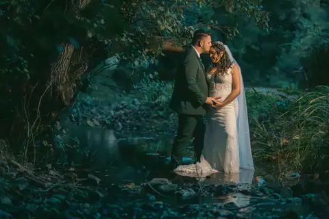 The couple embracing by the water, in the shade of a tree
